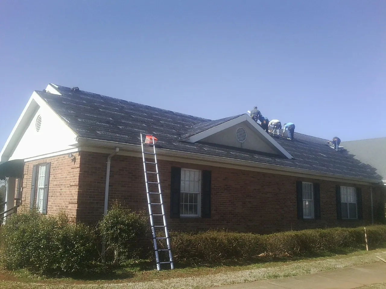 Crew tearing shingles off a brick ranch roof with a ladder set — Emergency Roof Repair work by our crew in Shutesbury