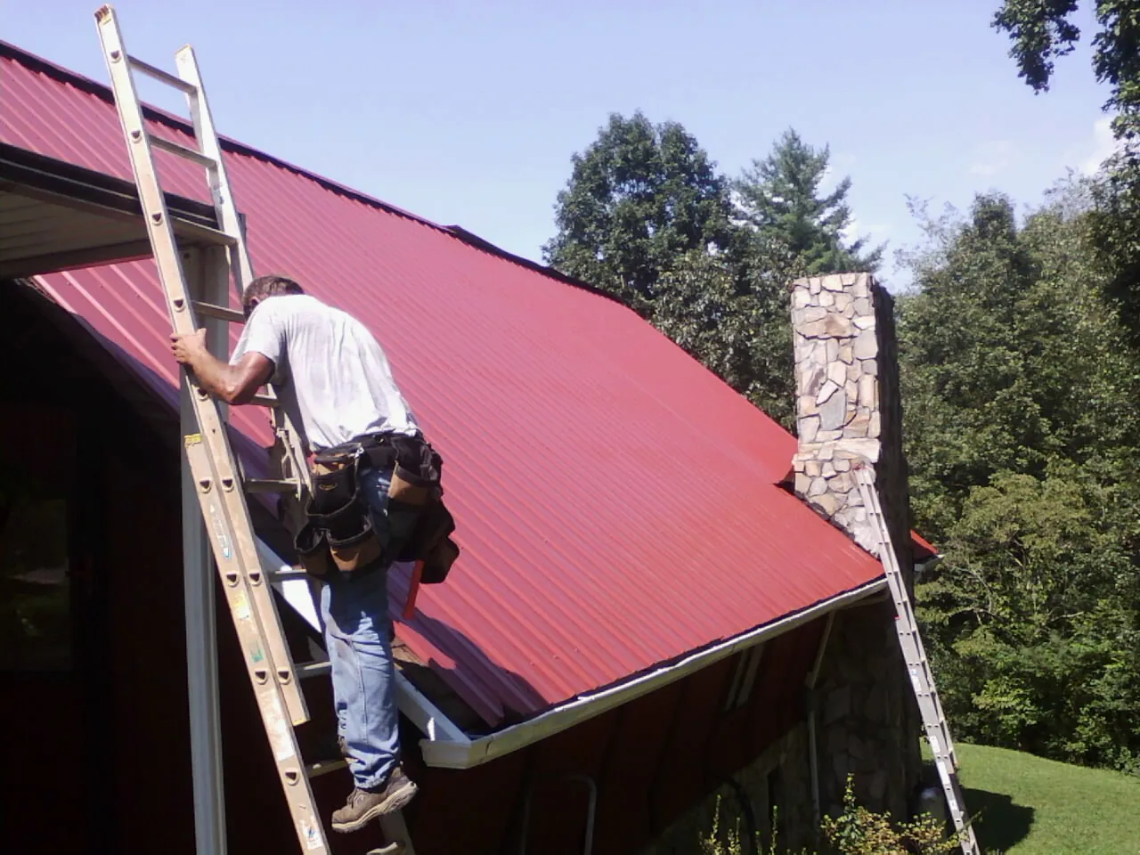 Roofer on a ladder beside a red metal roof and stone chimney — Soffit & Fascia Repair by Roofing Brothers in Shutesbury